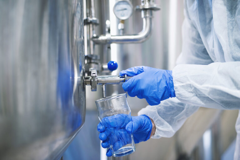 Close up view of hands in protective gloves opening tap valve and filling glass with samples from industrial machine reservoir